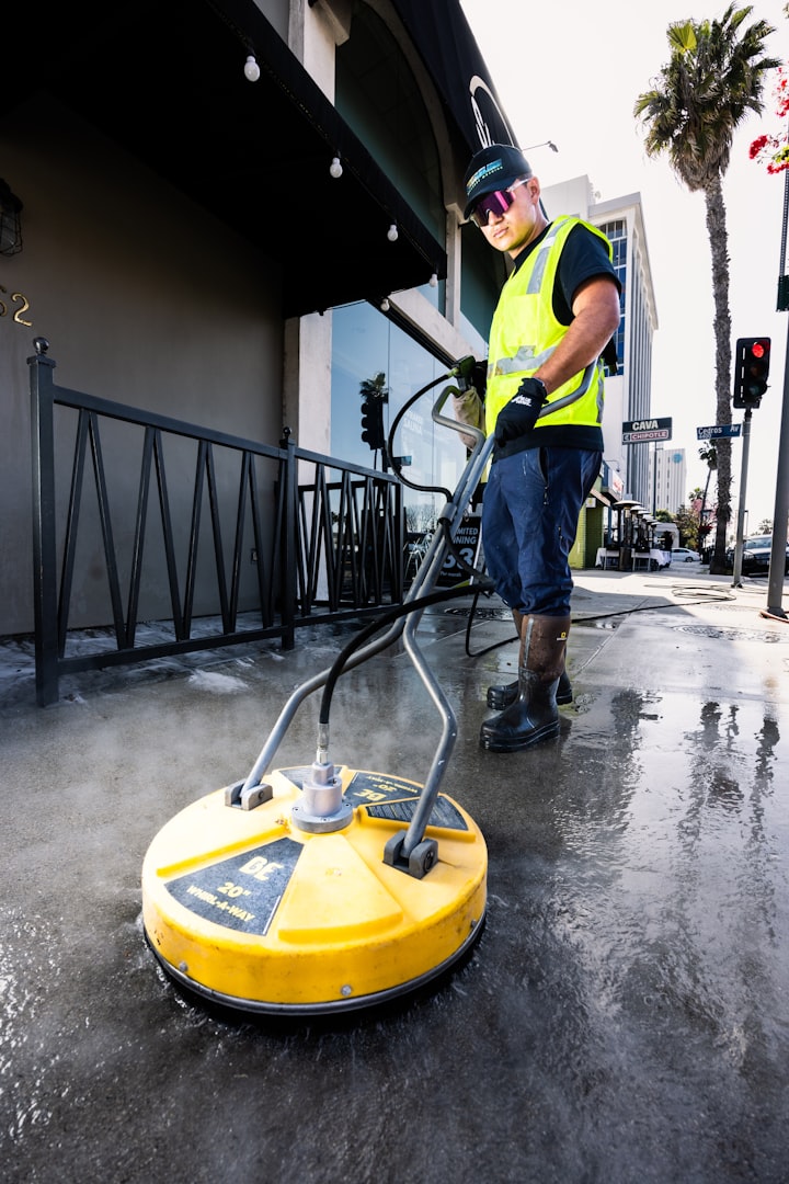 A person in overalls is using a pressure washer to clean a stone patio, with a lounge chair and neatly trimmed hedges visible in the background.